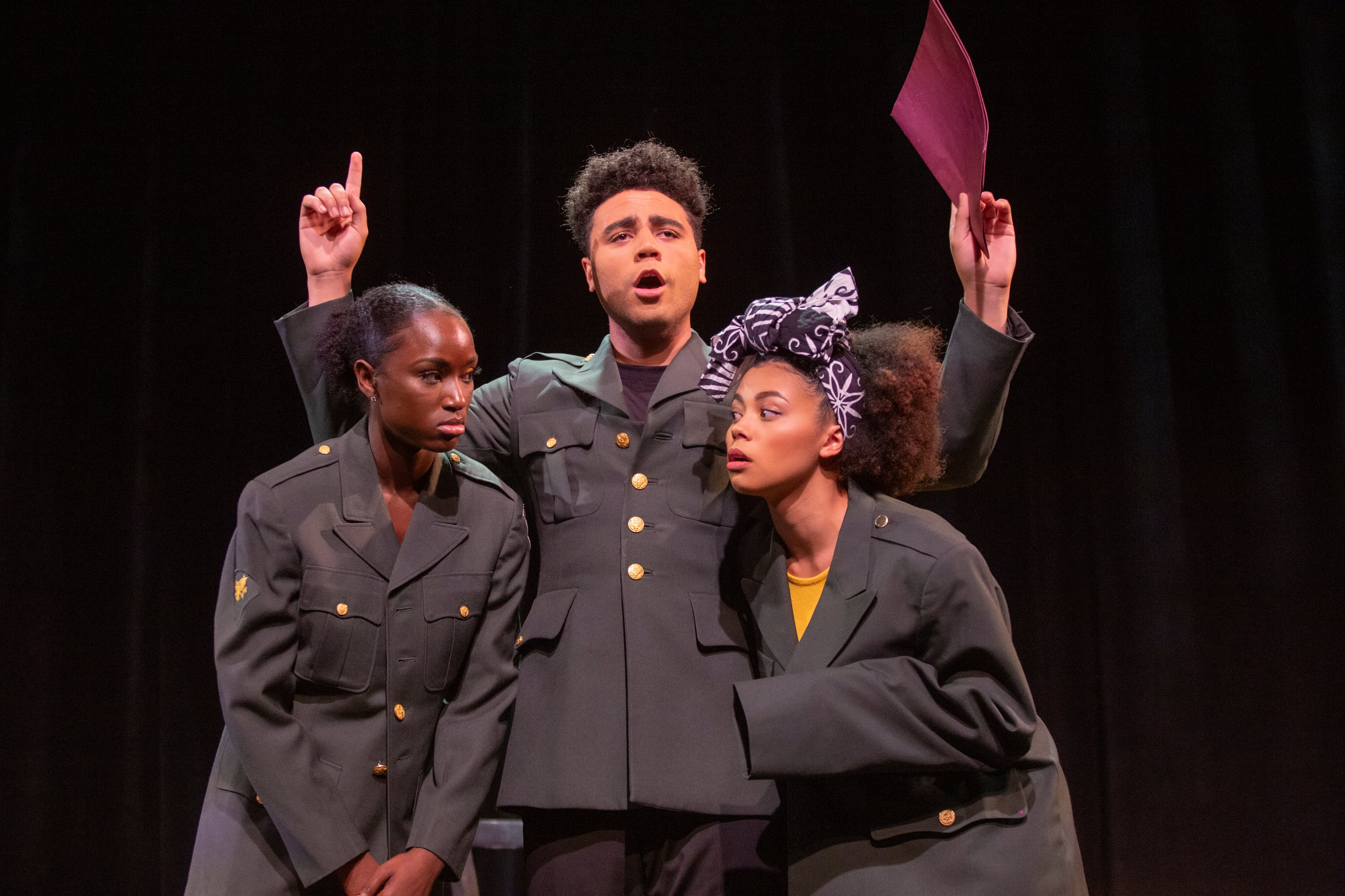 An actor holds a red folder in hand as two of his compatriots in matching uniforms glare at one another 