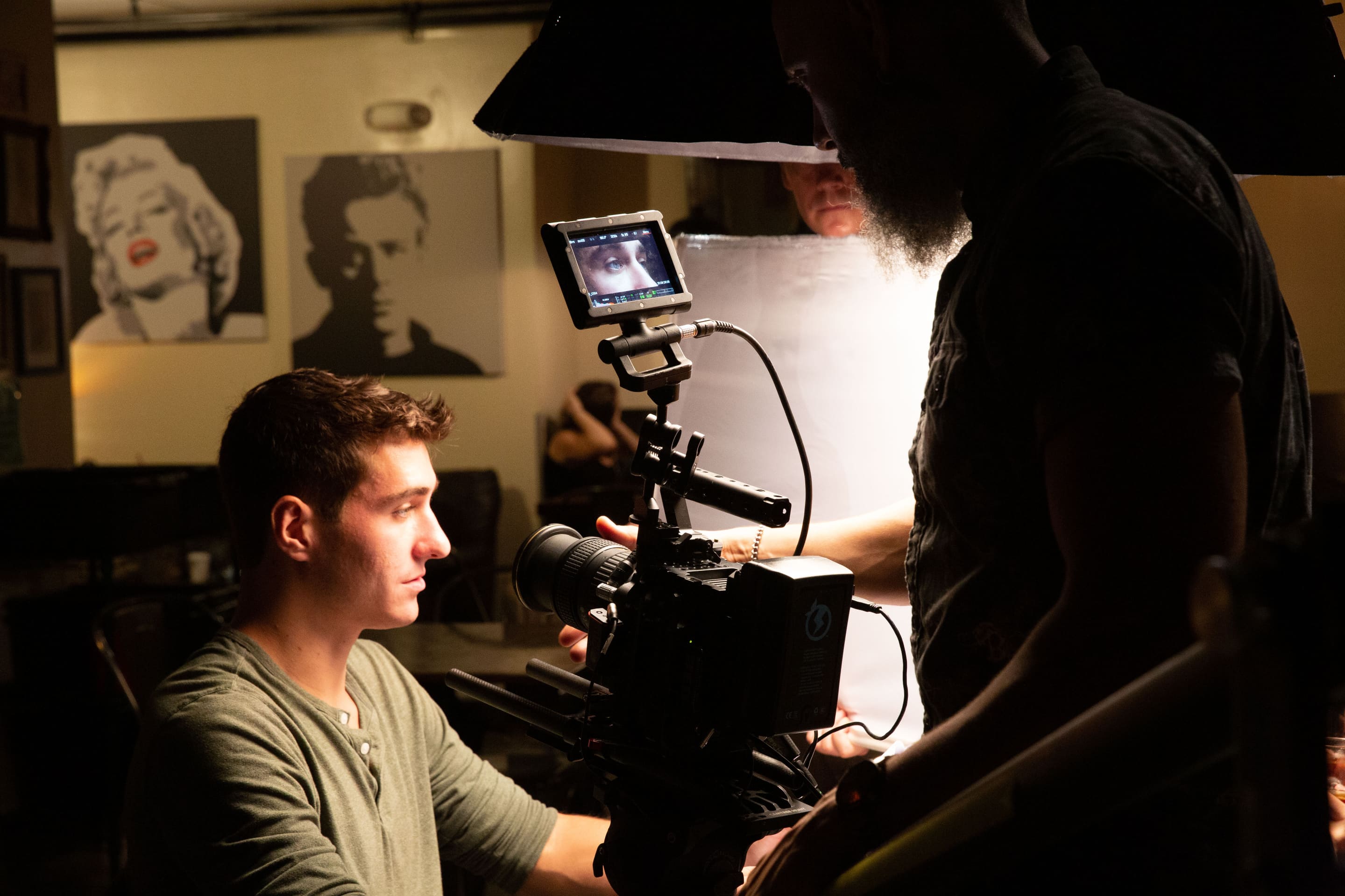 A sitting actor looks, unsmiling, across a table as cameraman zooms in on a shot focusing on his eyes