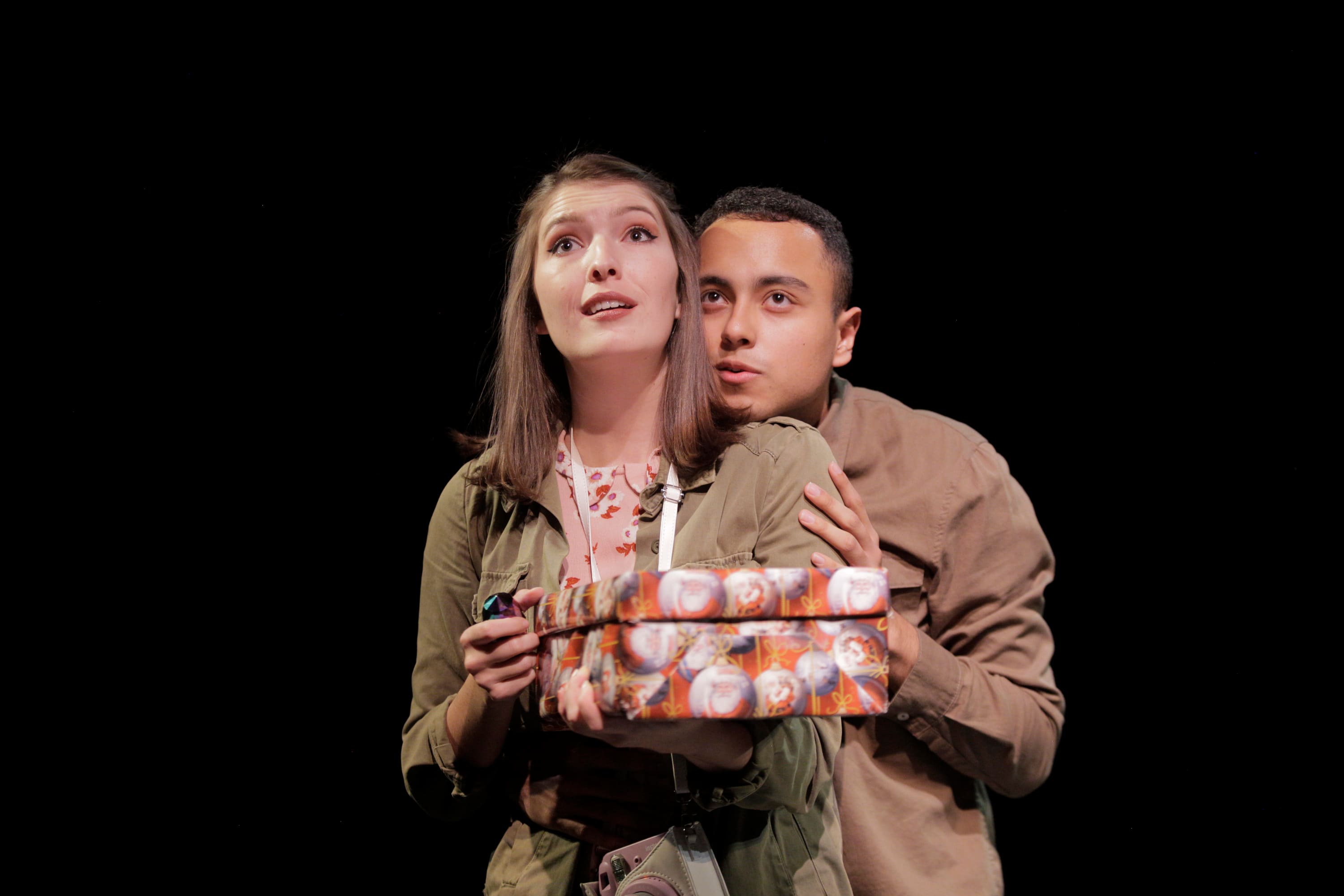 Two actors share a scene, one of them holds a gift wrapped box looking upwards as the other braces them from behind