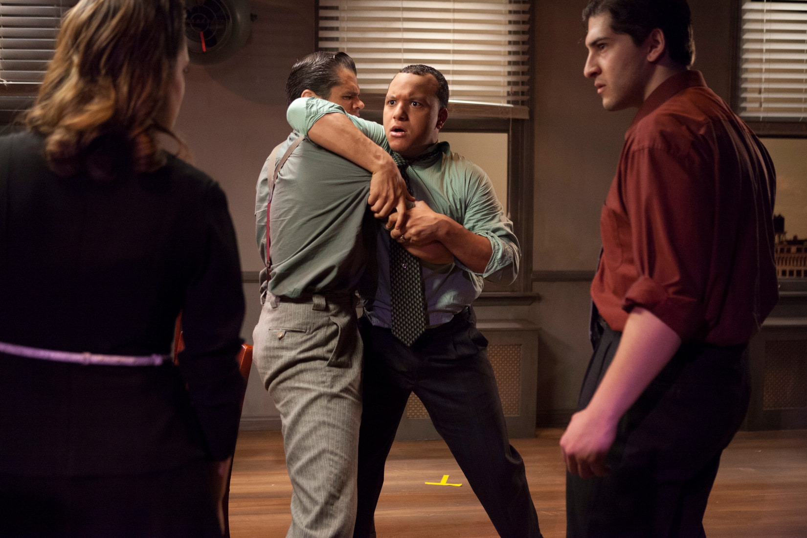 A dramatic scene of four, one man holds back another who is glaring enraged in the direction of an actor or potentially off-stage