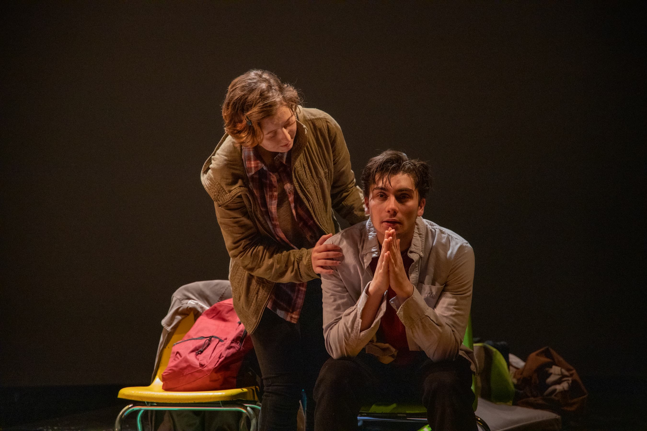 Two actors on stage: one sitting wide-eyed with hands praying as the other consoles him.