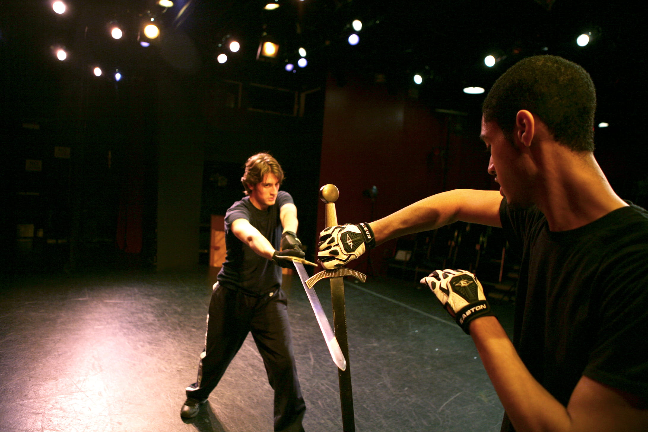 Two actors engaged in stage combat with swords. The individual closer to the camera parries a swing from the other