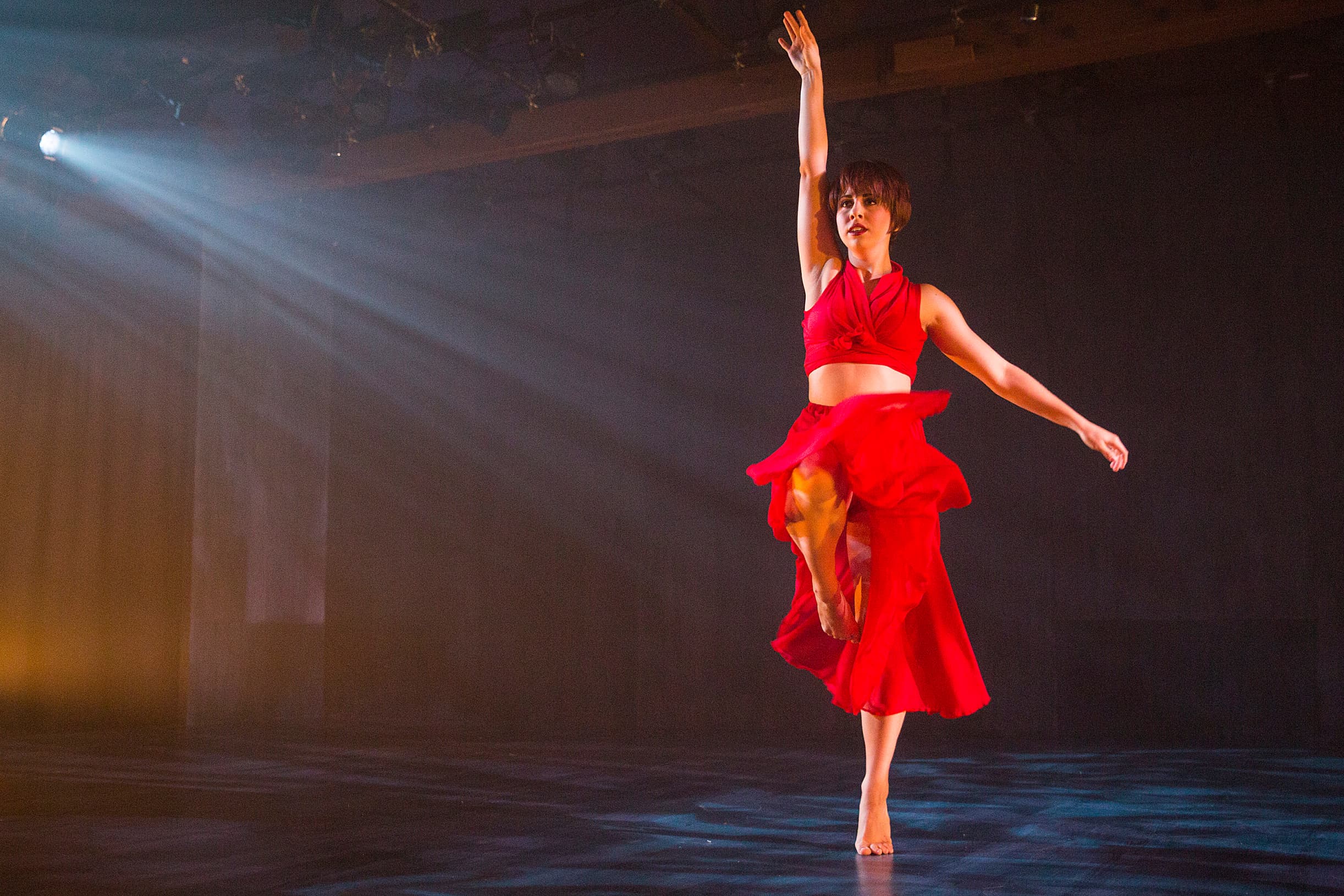 A dancer in a flowing red outfit strikes a pose on one leg with her right arm outstretched skywards