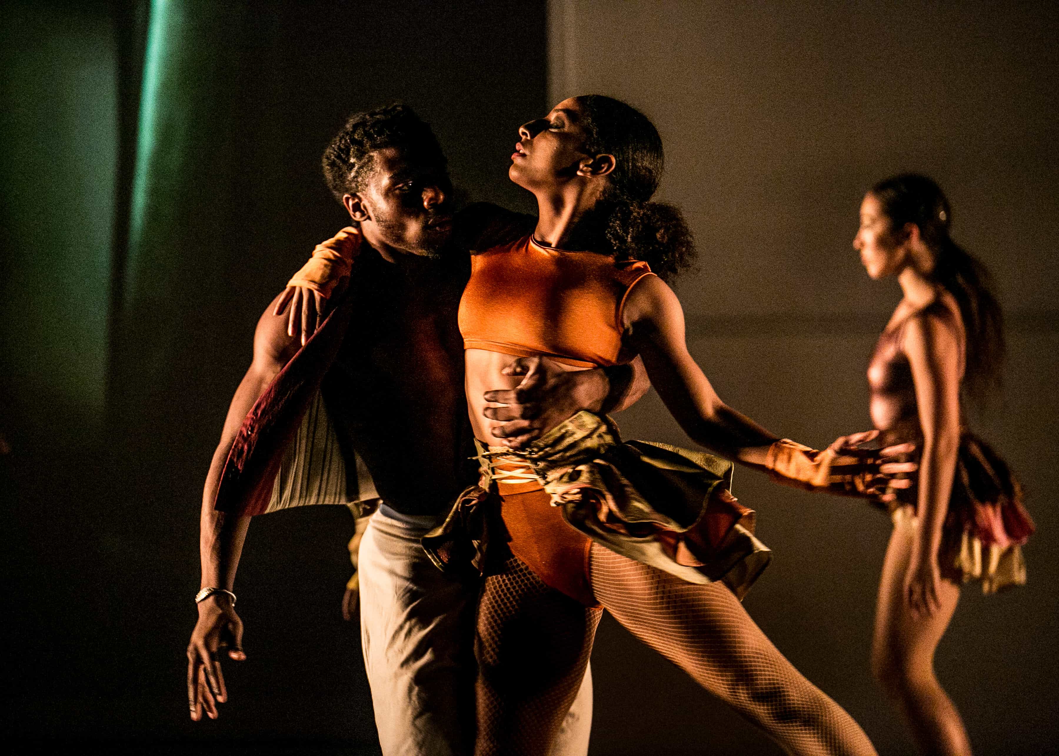 A dancer moves into the embracing support of another while a third dancer walks past in the background