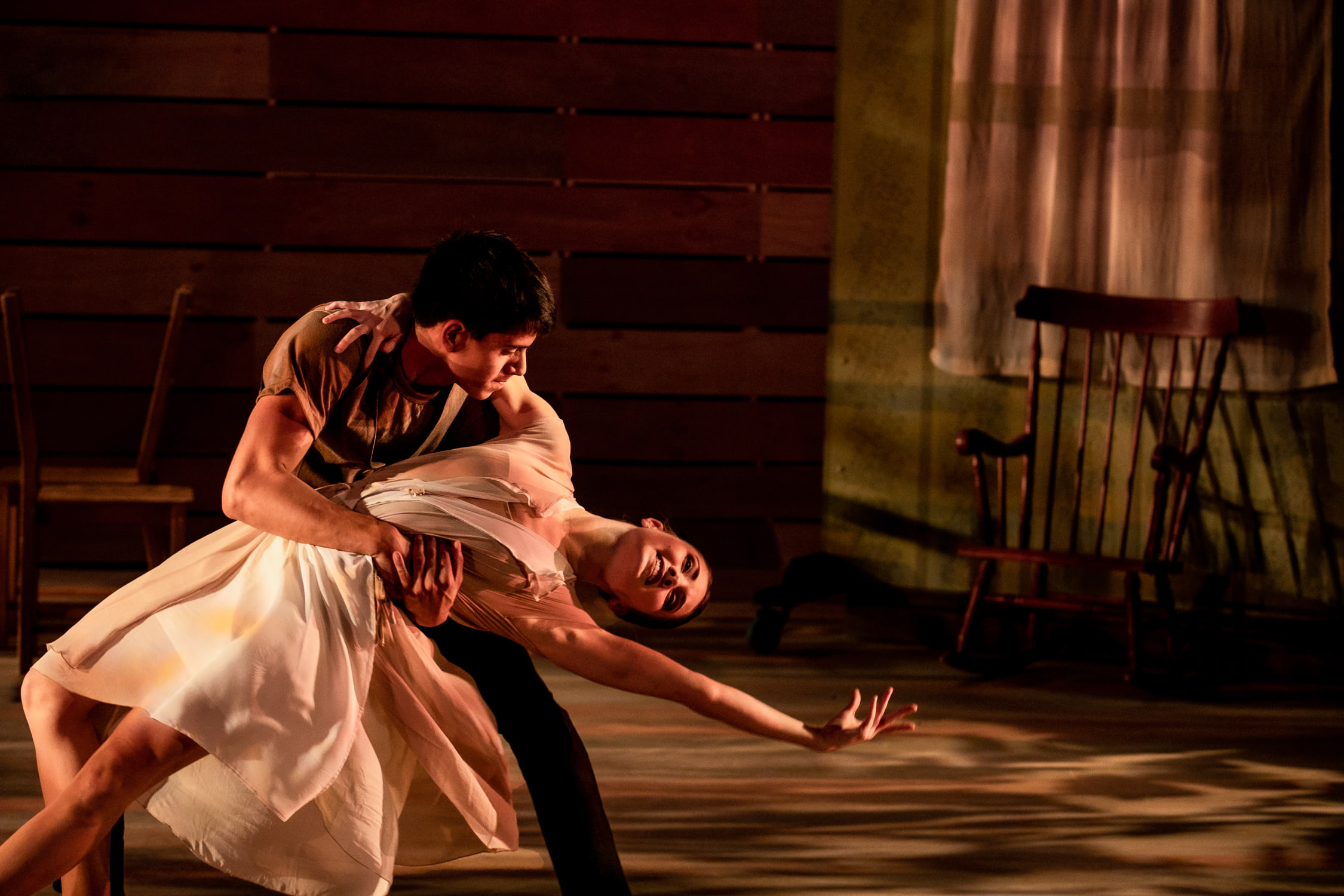 A dancer in a long white dress dips backwards with the assistance of another's embrace