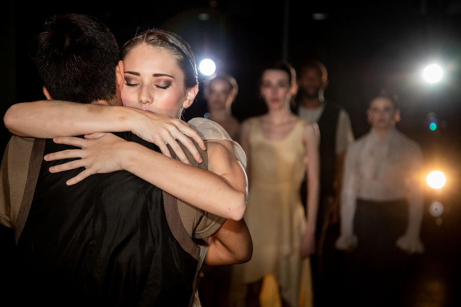 Two performers hug in the foreground of the stage