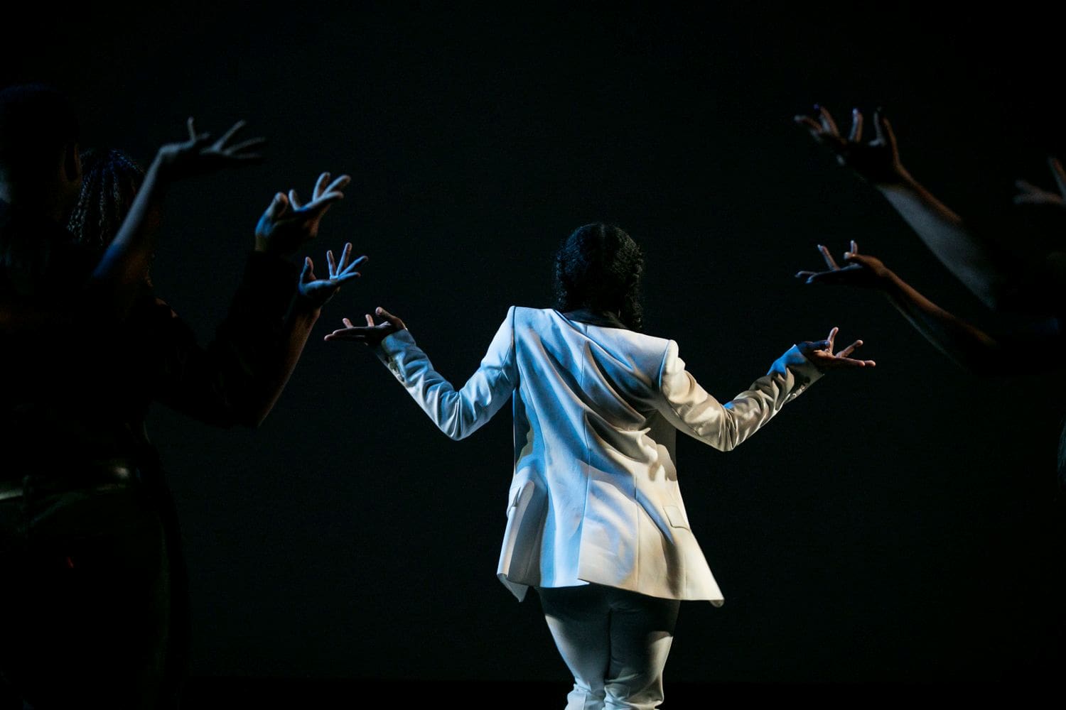 A dancer faces away from the stage center stage with arms raised