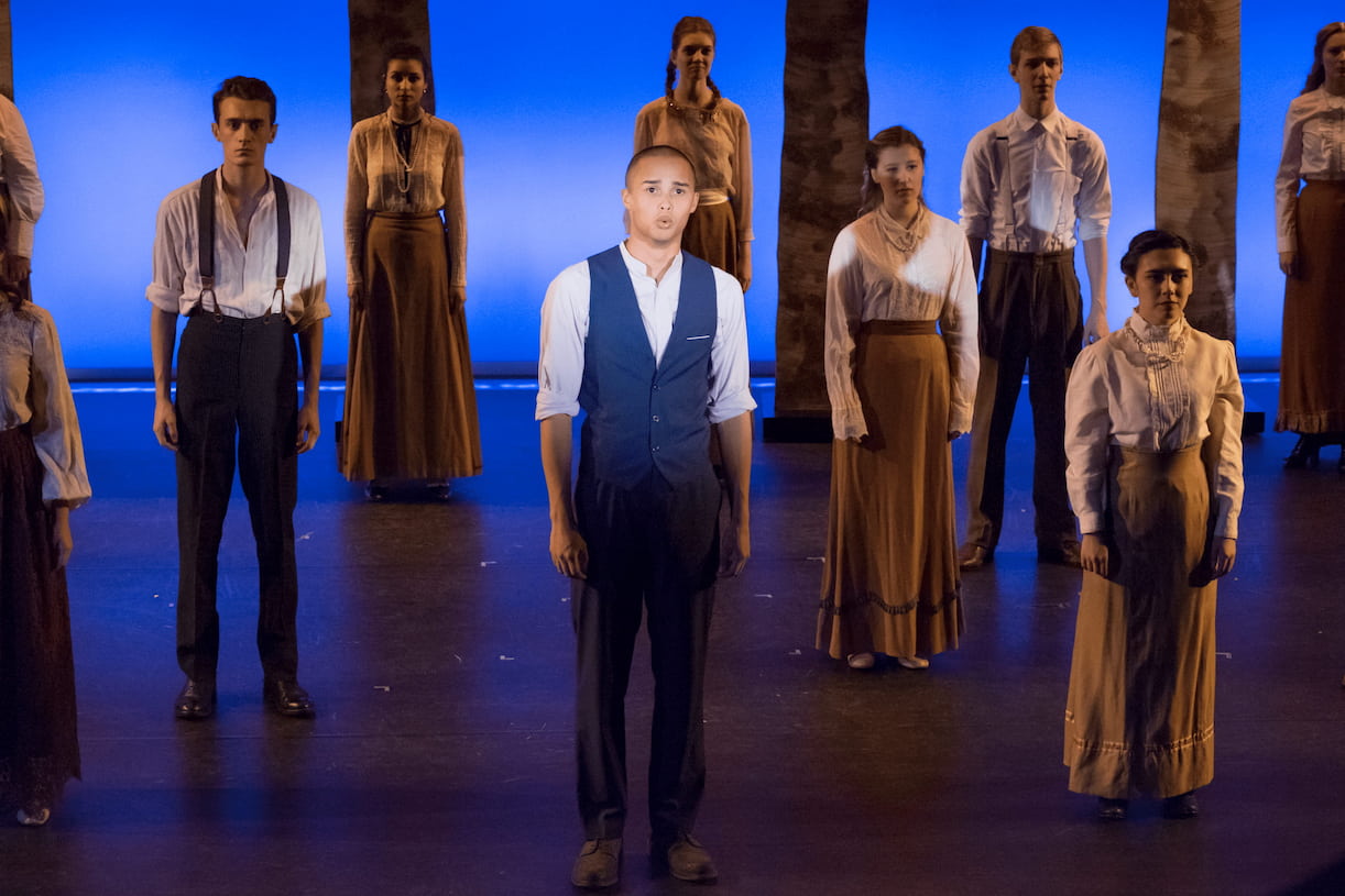 A performer sings a solo as solemn cast, evenly spread about a stage remain silent