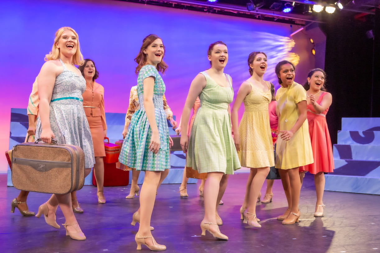 A group of performers in a variety of pastel-colored dresses sing in unison