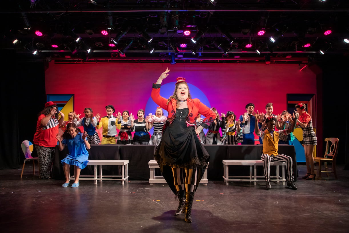 A vaudevillian actress takes center-stage singing while the rest of the cast, both hands raised, sing backup