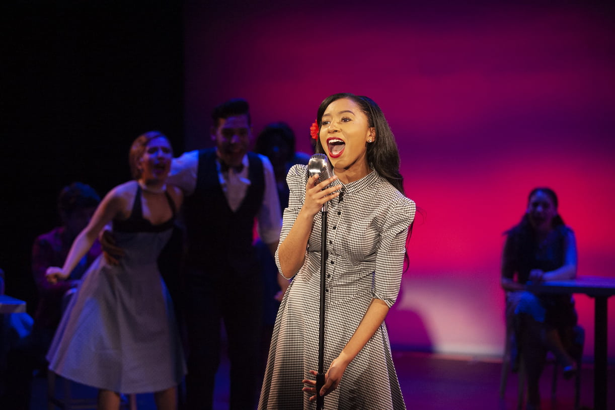 A singer in a black and white patterned dress and a red flower in her hair sings into a stand microphone