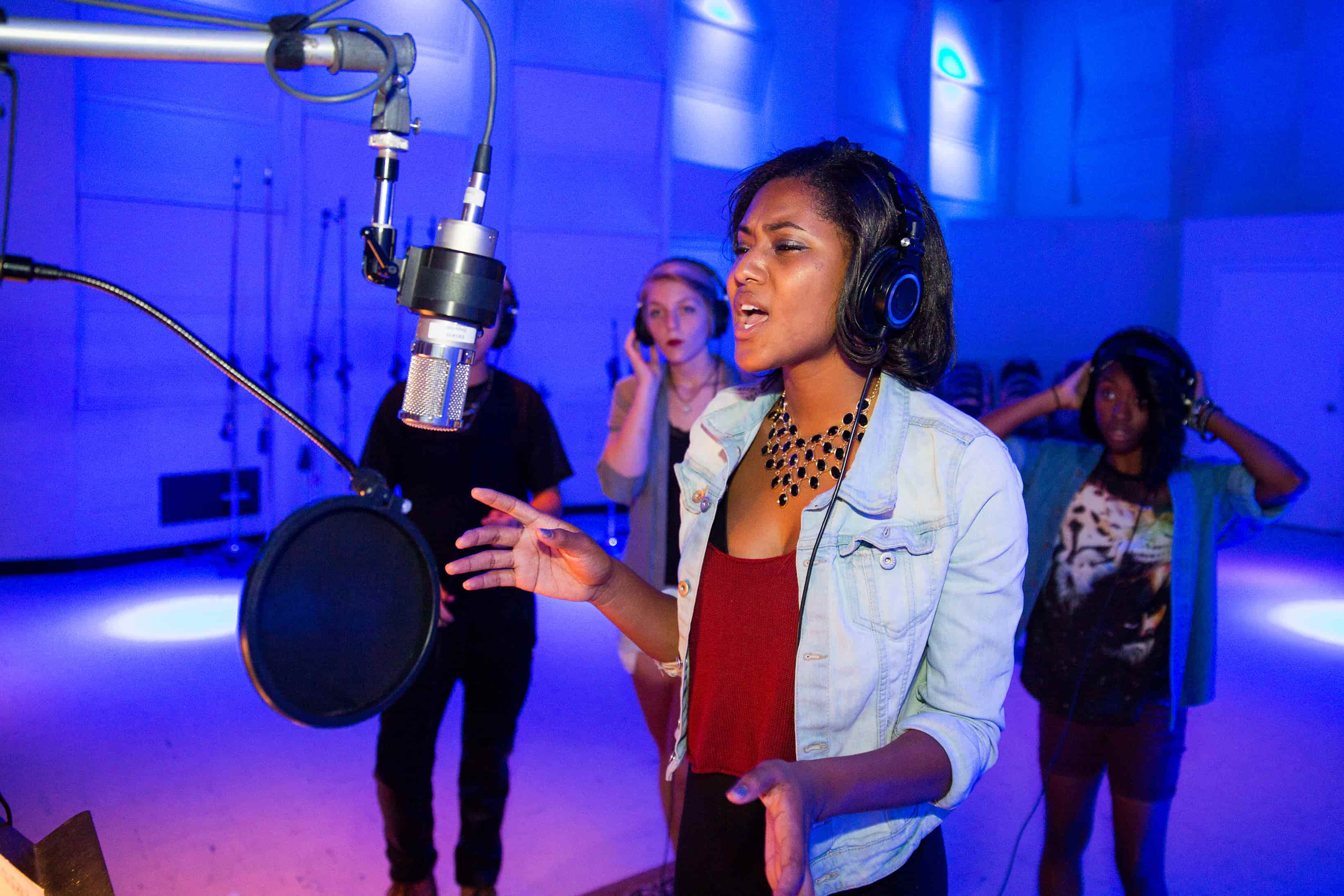 A vocalist performing in front of a recording microphone in a large studio alongside a group of individuals.